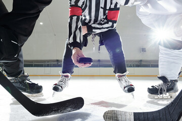 Hockey referee with puck standing on ice rink between two players with sticks