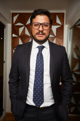A young Latino businessman wearing glasses and standing in a dark suit and tie