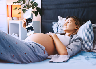 Pregnant woman looking at ultrasound image relaxing in bed at home. Expectant mother waiting and preparing for baby.