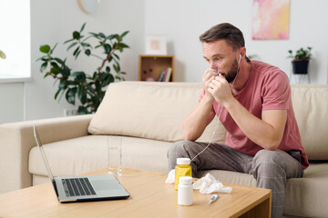 Young sick patient in earphones blowing his nose while talking to online doctor