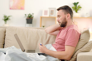 Side view of young sick man with thermometer sitting on couch under blanket