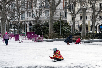 Una niña está sentada en el suelo jugando con la nieve. Lleva un abrigo de invierno y un gorro de...