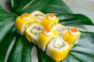 Side view of Japanese sushi roll with unagi eel, cream cheese, avocado wrapped in mango slices served with red caviar on top. exotic monstera leaf on gray stone background. food art concept

