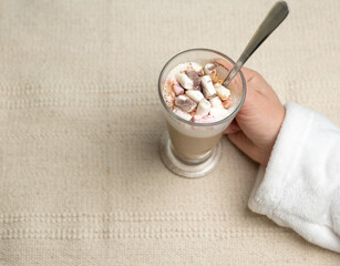 cappuccino in a transparent glass with marshmallows and cocoa powder in female hands on a textured beige fabric