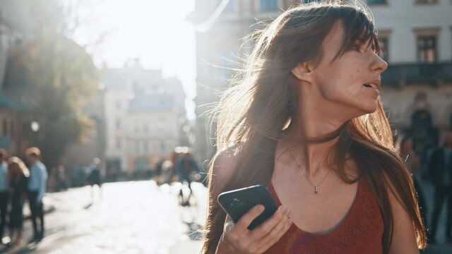 Motion camera view of cheerful caucasian woman with vitiligo disease looking at the google maps at mobile phone while walking in street with ancient buildings at the background. Women looking around