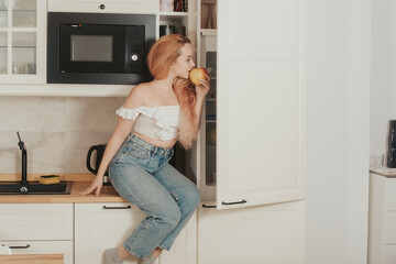 A woman is sitting at home in the kitchen near the refrigerator. A girl with a good figure looks in the refrigerator, healthy food, diet. The girl sits on the table and eats an apple