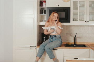 The woman is sitting at home in the kitchen. A beautiful girl drinks tea or coffee sits on the table and speaks on the phone. A student orders food at home.