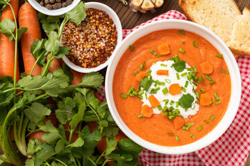 Carrot soup of freshly ground carrots with herbs and sour cream on a wooden background.