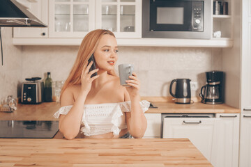 A beautiful girl is sitting in the kitchen drinking tea or coffee and looking at the phone. Online shopping, delivery from food stores. The girl speaks on a smart phone and smiles