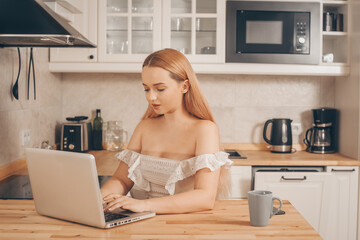 Fototapeta na wymiar A beautiful girl sits at home in the kitchen and works at a laptop. There is a cup of coffee or tea nearby. Freelance woman, work from home, covid restrictions.