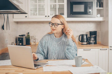 Obraz na płótnie Canvas A woman works from home using a laptop. A beautiful blonde in a sweater and glasses, there is a mug and papers on the table. Home schooling, work online. A student with headphones speaks by video 