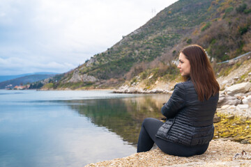 Fototapeta na wymiar Young brunette woman in jacket sits and reflects on rocky shore of calm sea against background of mountains in peaceful atmosphere. Idyllic walk alone.