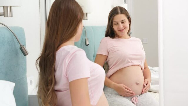 Beautiful smiling pregnant woman sitting on bed and looking in her reflection at big mirror. Beautiful pregnancy and anticipation of baby.