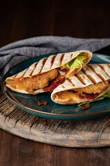 Close up of two grilled panini wraps filled with breaded chicken, bacon and cheese against a dark background.