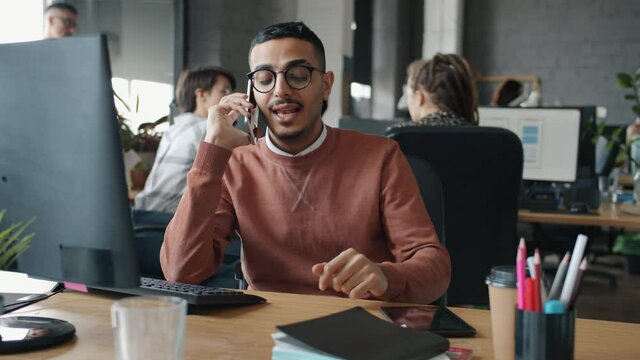 Serious Arab man is speaking on mobile phone and looking at computer screen working in open plan office with people in background. Communication and technology concept.