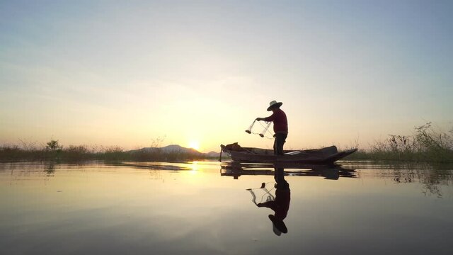 Footage B roll of Silhouette fisherman throwing fishing net during sunset with boats at the lake. Traditional culture of Thailand. Concept Fisherman's life style.