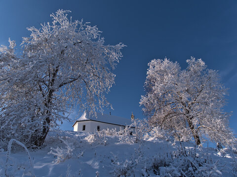 Low angle view of the top of Kornb&uuml;hl hill near Burladingen, Swabian Alb, Germany with snow-covered historic chapel Salmendinger Kapelle in winter season with frozen trees on sunny day.