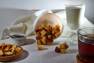 Square toasted pieces of homemade delicious rusk, hardtack, Dryasdust, zwieback, Liquid honey in a saucer, glass of milk and black tee in a cap on a white tablecloth.