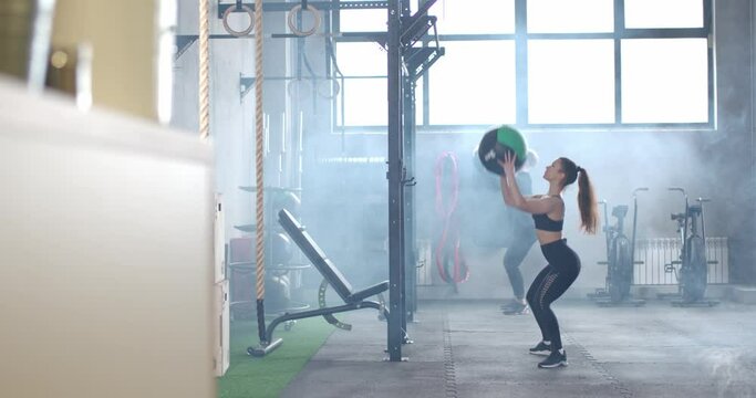 Crossfit training, young athletes perform squat and throw balls, man and female trains in the gym.