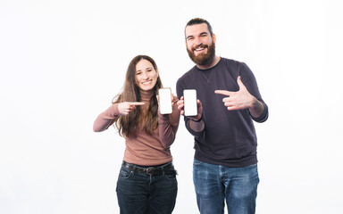 Smiling young couple standing over white background and pointing at smartphone screens.