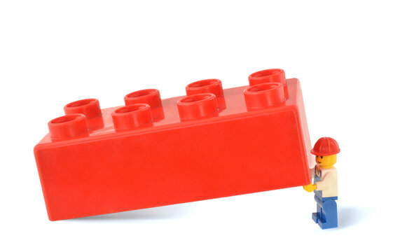Editorial illustrative image with concept of heavy work. Lego minigifure in blue overall gets up a heavy big plastic block or red brick.