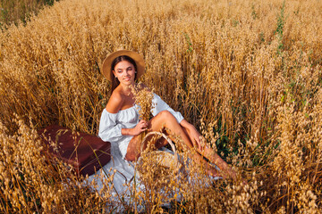 Young beautiful woman with straw hat sitting at golden oat field near the basket with ears of oats.