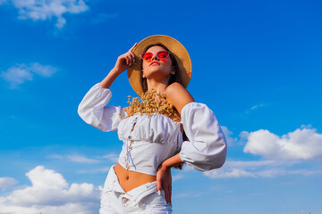 Fashion portrait of a young beautiful woman with straw hat and pink sunglasses with oats in her blouse posing on a blue sky background.