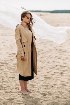 Female loneliness. Fall nostalgia. Despair sorrow. Art portrait of thoughtful depressed woman in beige trench coat with flying away hair in cold wind standing barefoot on sandy beach.