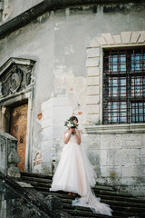 Bride in wedding dress with a bouquet of flowers stand on stairs near wall ancient architecture, old building.