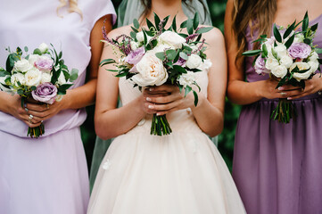 The bride and bridesmaids in an elegant violet dress is standing and holding hand bouquets of...