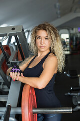 Fototapeta na wymiar Woman with long blonde curly hair - portrait of a fitness trainer in the gym. Sports form, healthy lifestyle, muscle building, weight loss. Sports wear, gloves without fingers. Beauty and confidence