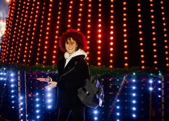 Curly girl with the backpack walks in the night city on a festive night, the lights of city are new year.