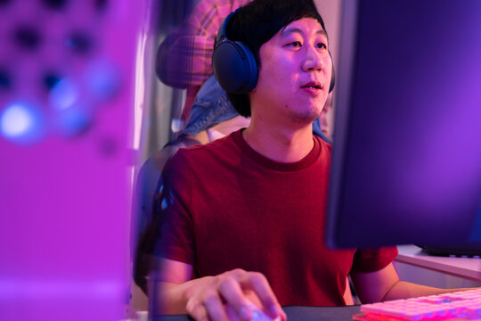Young asian male working from home in front of a professional computer station using a pair of headphone. Young boy playing video game from his studio. Gamer and technology lifestyle concept.