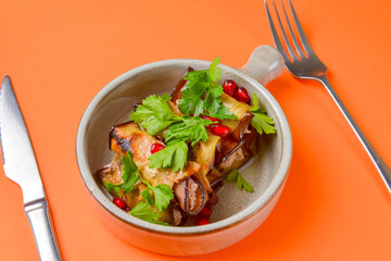 Egg-plant rolls with meat and pomegranate seeds served in a bowl with knife and fork over orange background.