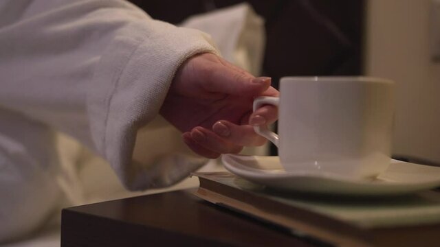 White robe, lying soft bed hotel room, hand takes cup from bedside table book. Travel hotel relaxation, unforgettable weekend. Nice good morning. Delicious drink after wellness treatments, warm shower