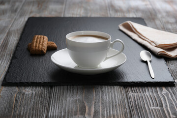 White cup of cappuccino, small spoon, cookies on a black textured stone plate, wooden textured table. White foam.