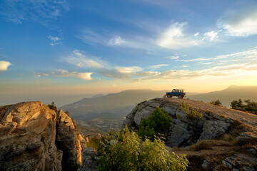 Jeep near a steep cliff against the background of mountains and the setting sun in the blue sky