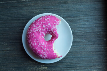 Top view to bright pink donut on white round plate bitten off twice. Aged wooden background....