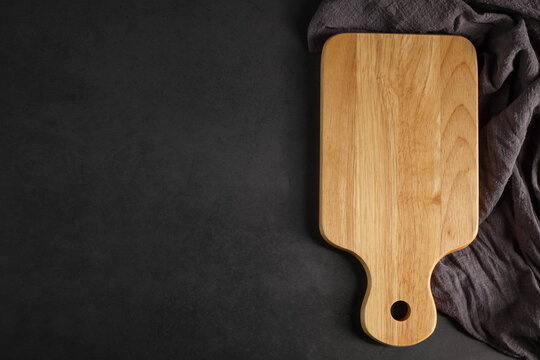 Top View Above Of Wooden Chopping Board With Apron On Dark Black Table Background. Empty Wood Cutting Board With Handle And Hole For Hanging With Copy Space.