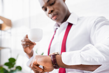 Wristwatch on arm of african american businessman with coffee cup on blurred background at home