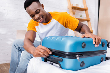 Positive african american man locking suitcase on blurred foreground on bed