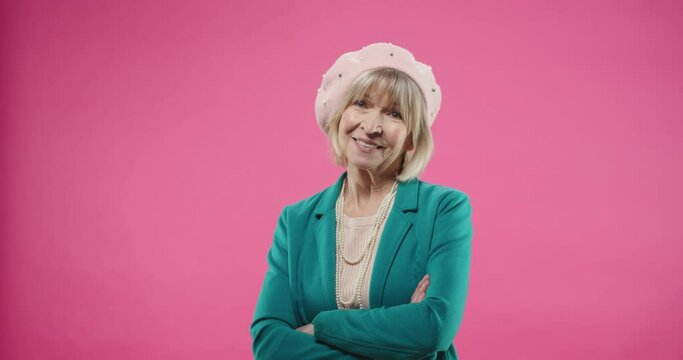 Portrait of happy smiling Caucasian senior old female in green jacket and beret standing isolated on pink background in positive mood looking at camera with happy face expression alone