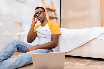 Tired african american man in eyeglasses sitting near laptop on blurred foreground in bedroom