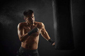 Fototapeta na wymiar male boxer in black boxing wraps punching in boxing bag on dark background with smoke