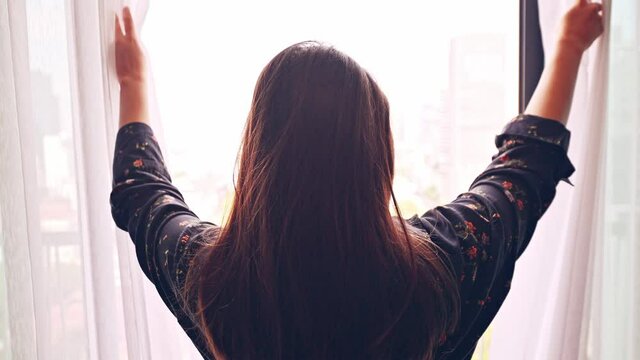 Lovely girl is awake and standing before window. Beautiful woman opening curtains and looking Cityscape view.