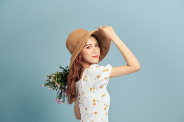 Close up portrait of an attractive young woman in summer dress and straw hat holding flower bouquet and looking over her shoulder isolated over blue background