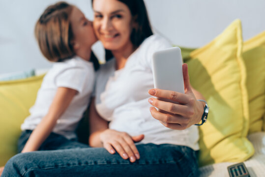 Daughter kissing smiling mother during selfie at home on blurred background