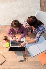 Top view of mother pointing with finger at laptop while sitting near daughter at desk with stationery