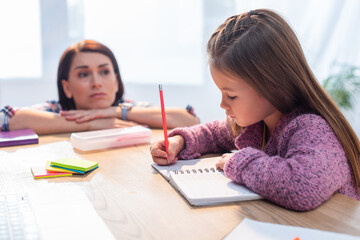 Upset mother looking at daughter writing in notebook at desk on blurred background