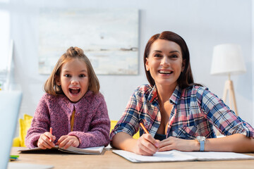 Smiling mother and excited daughter with open mouth looking at camera with blurred living room on background